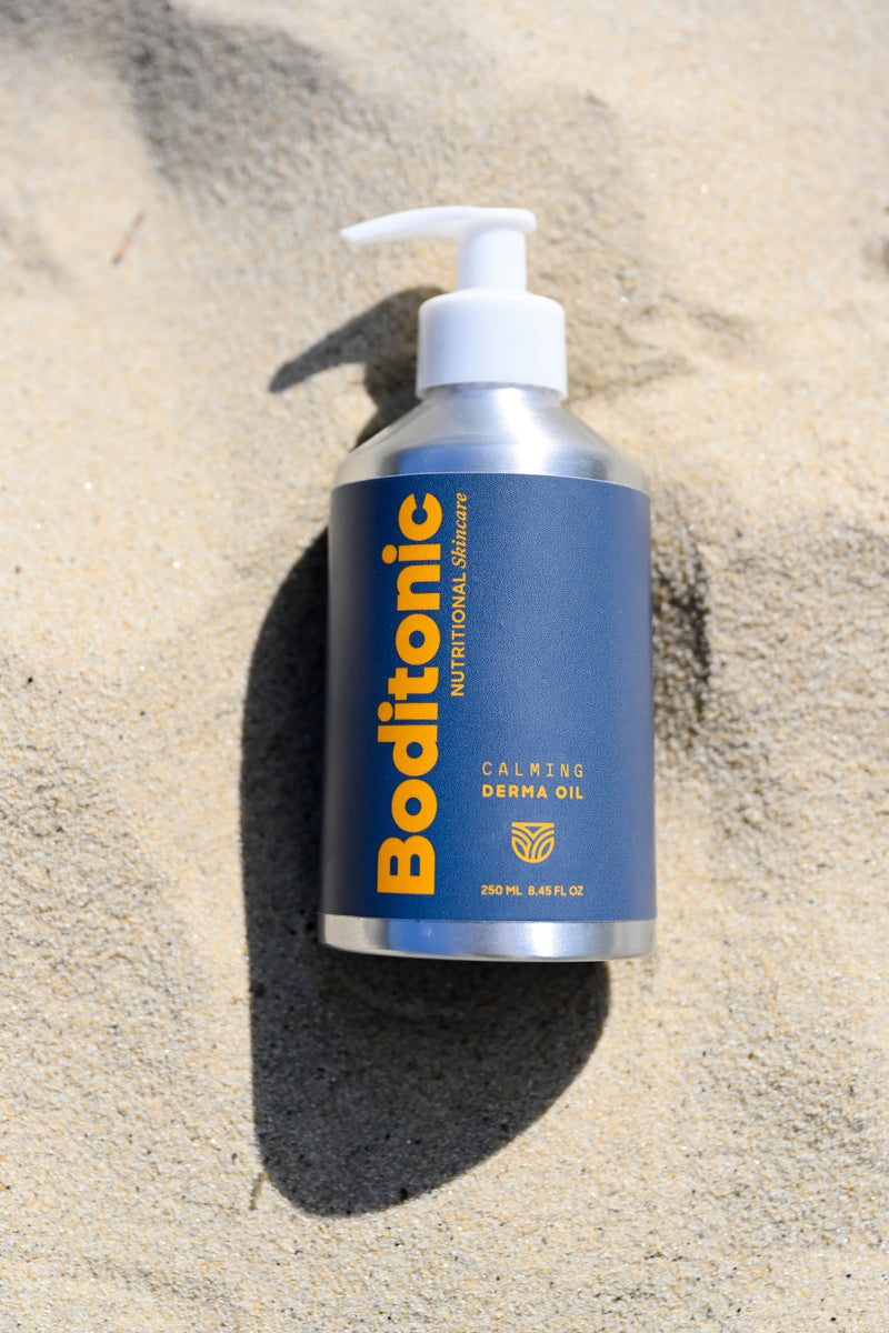 Boditonic Calming Derma Oil 250 ml bottle lying in warm beach sand in bright sunlight